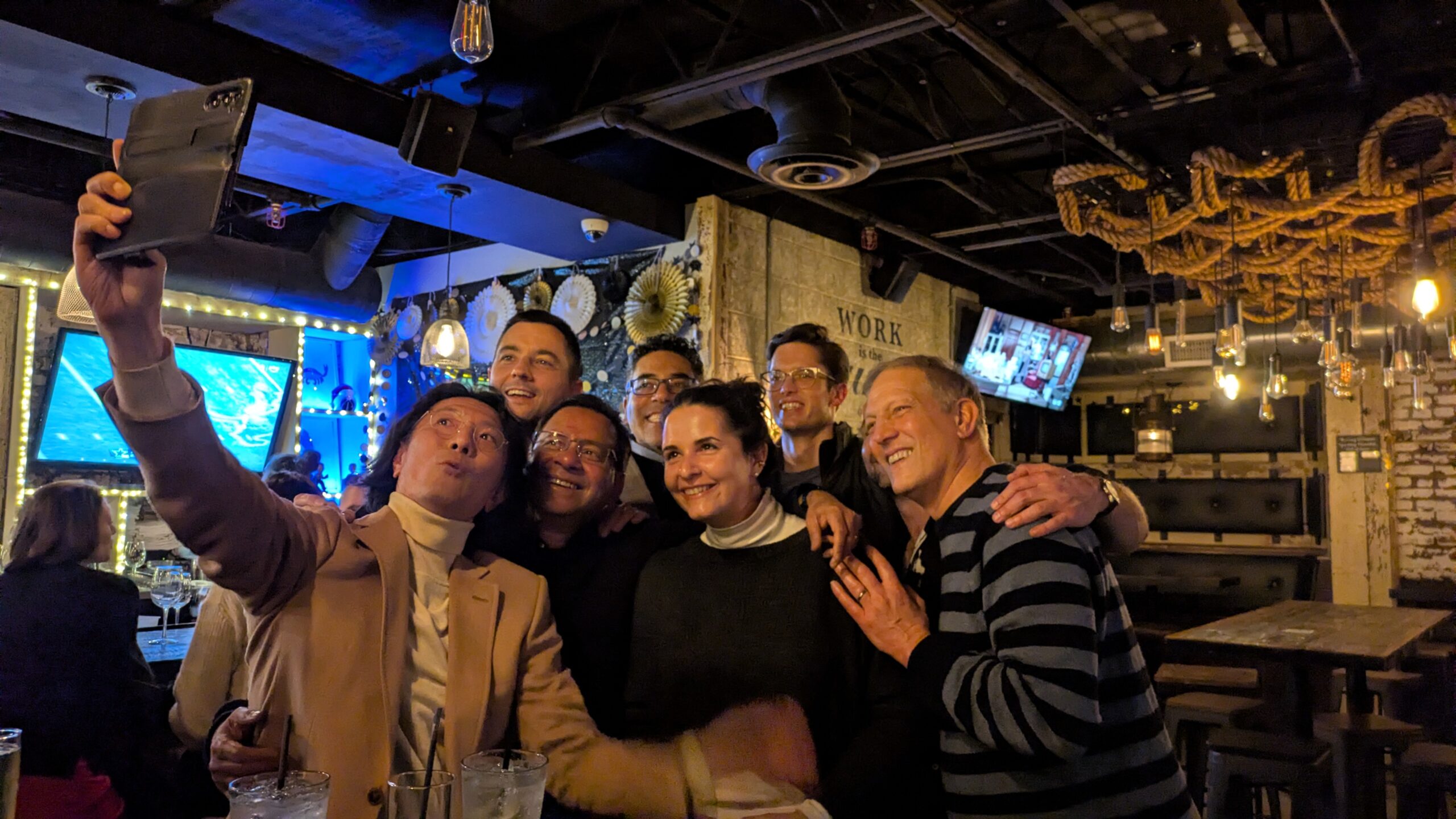 A group of six people pose for a selfie at a lively bar with drinks on the table, TVs in the background, and warm lighting.