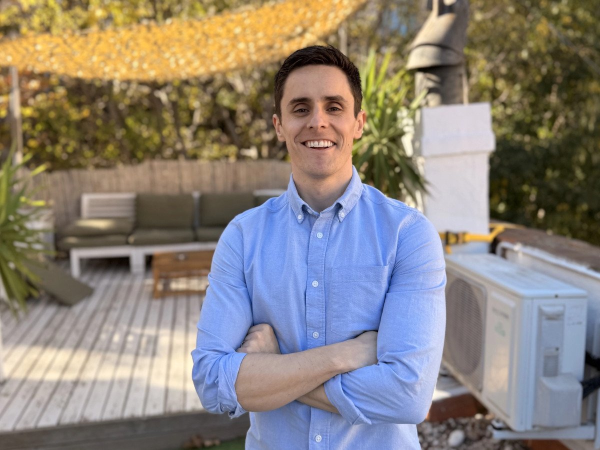 A man in a blue shirt stands in an office garden-type area. He is smiling and has his arms crossed.