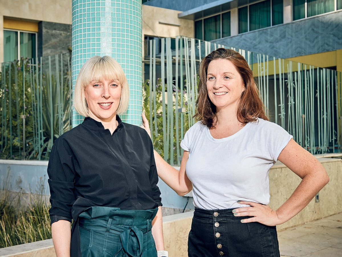 A woman with light blonde hair with fringes and a brunnete haired woman standing side by side.