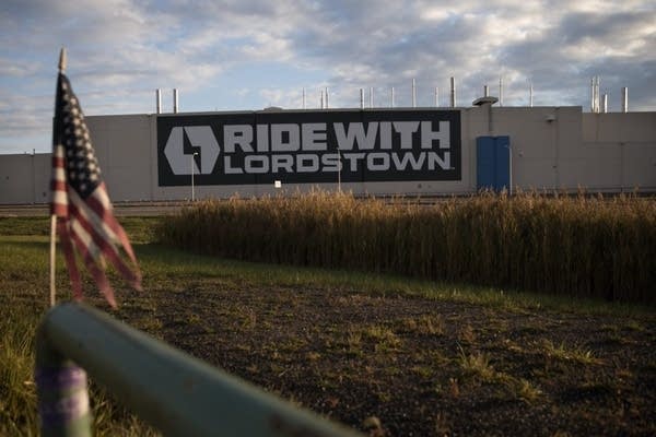 The Lordstown Motors factory in Lordstown Ohio. It's an industrial, white steel building with a massive electric logo and text that reads "Ride With Lordstown." It is seen behind grass and an American flag.
