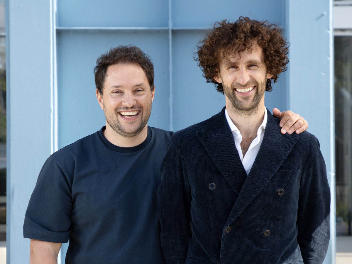 Two white men smile and pose for the camera in front of an office building.