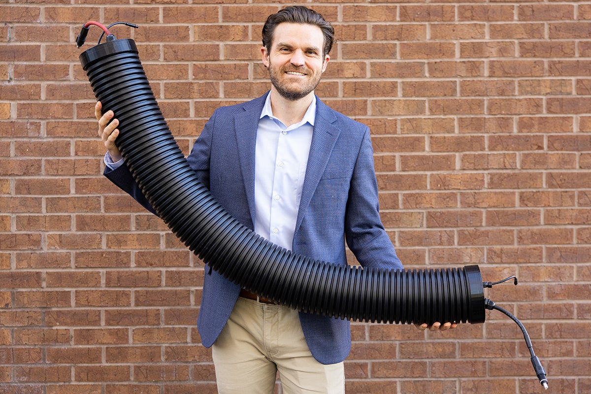 Man in blue professional jacket and khaki pants holds black tube with wires on ends while standing in front of brick wall