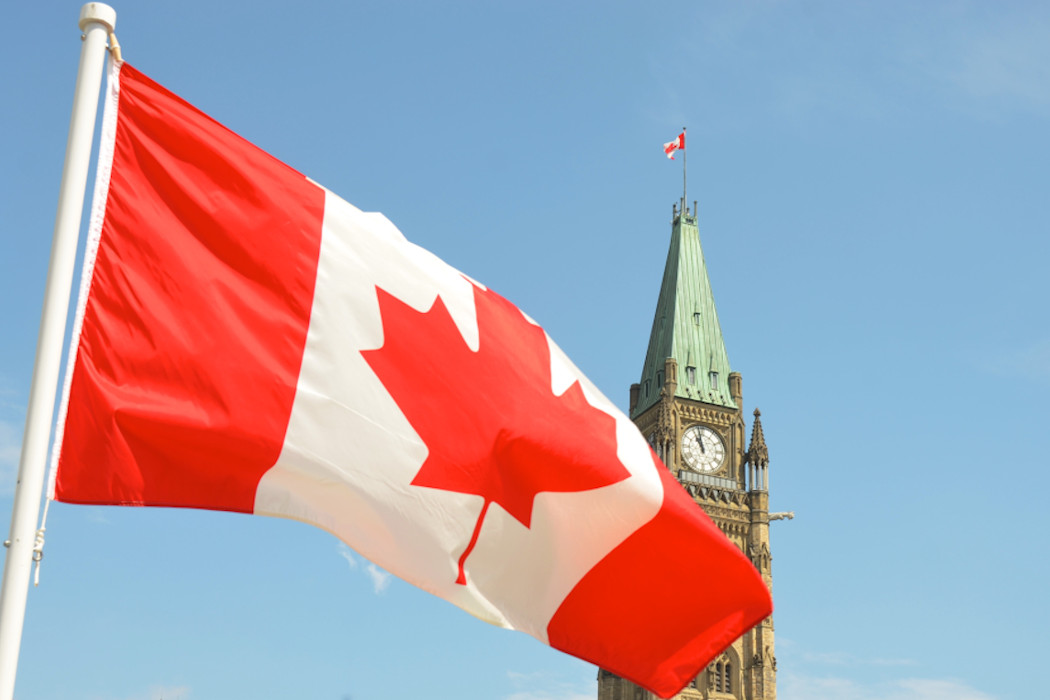 Canadian flag flies in the foreground over the parliament buildings in Ottawa on a sunny day