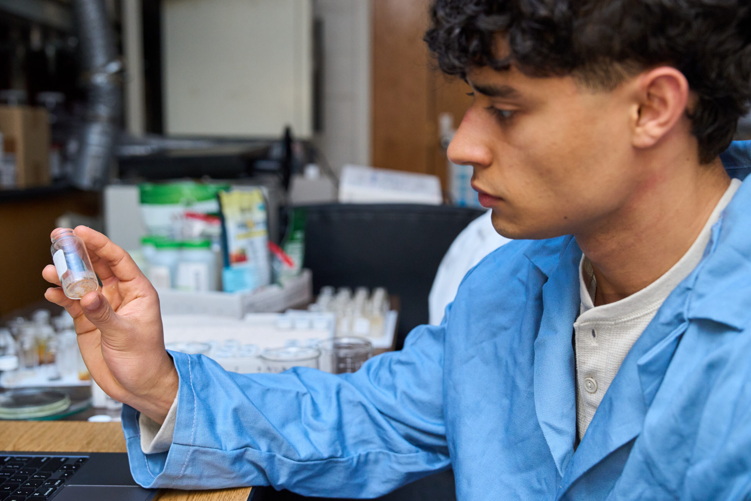 A man wearing a blue lab coat holds up a test tube with crystals in it while in a science lab.