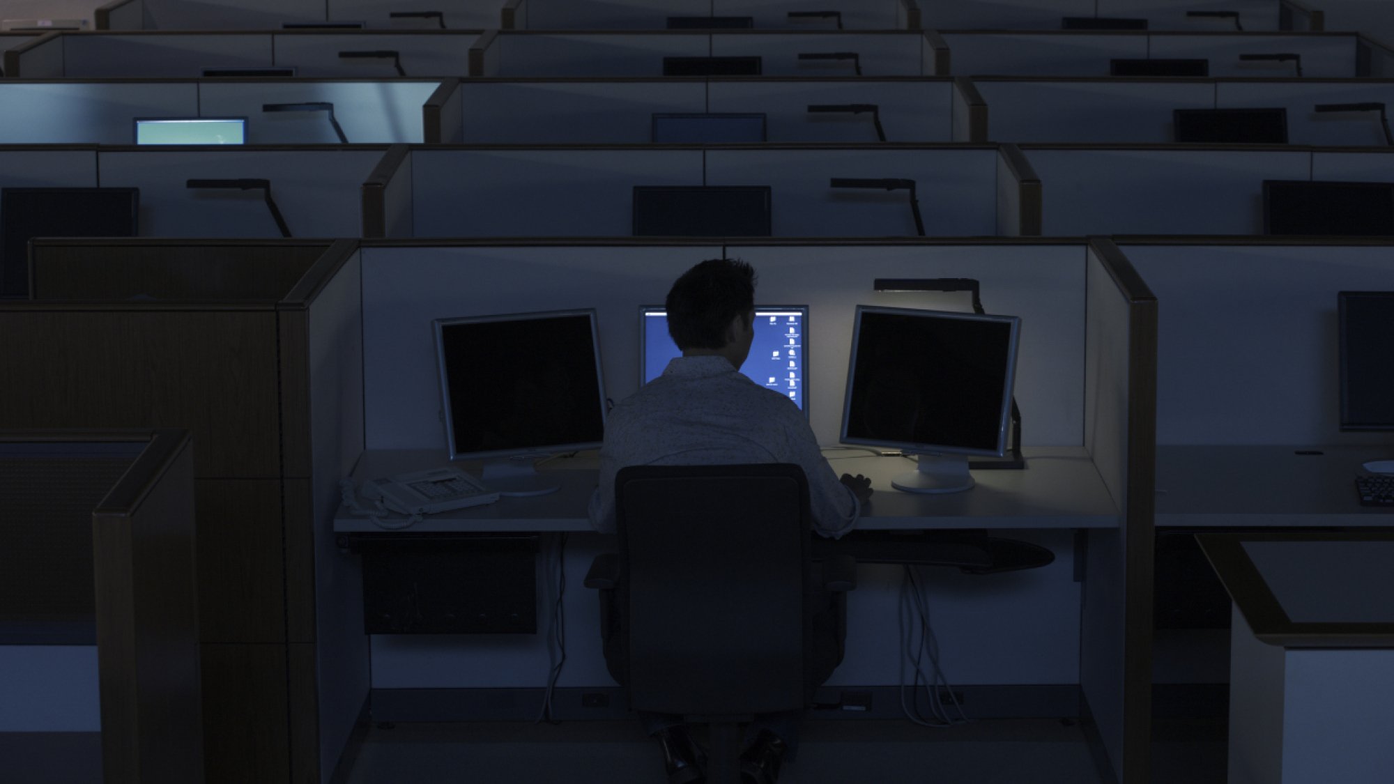 a lone worker in a dark call center sits in front of a computer