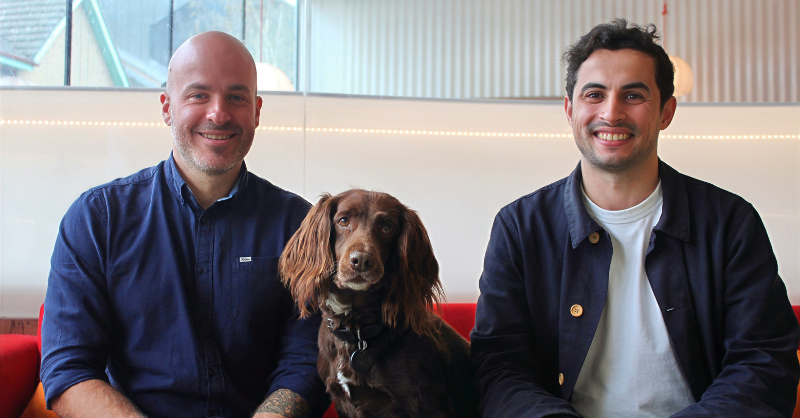 Two men sit together with a dog in between.