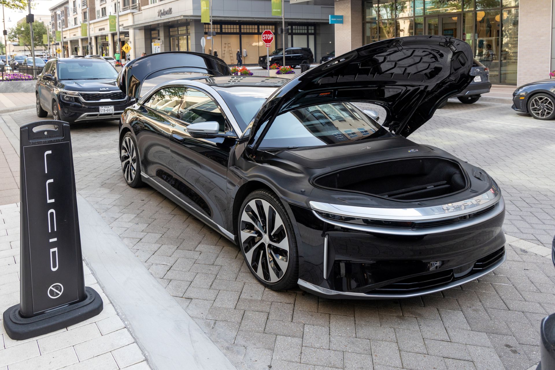 A black electric vehicle with its front trunk open is parked on a city street next to a Lucid Motors sign.