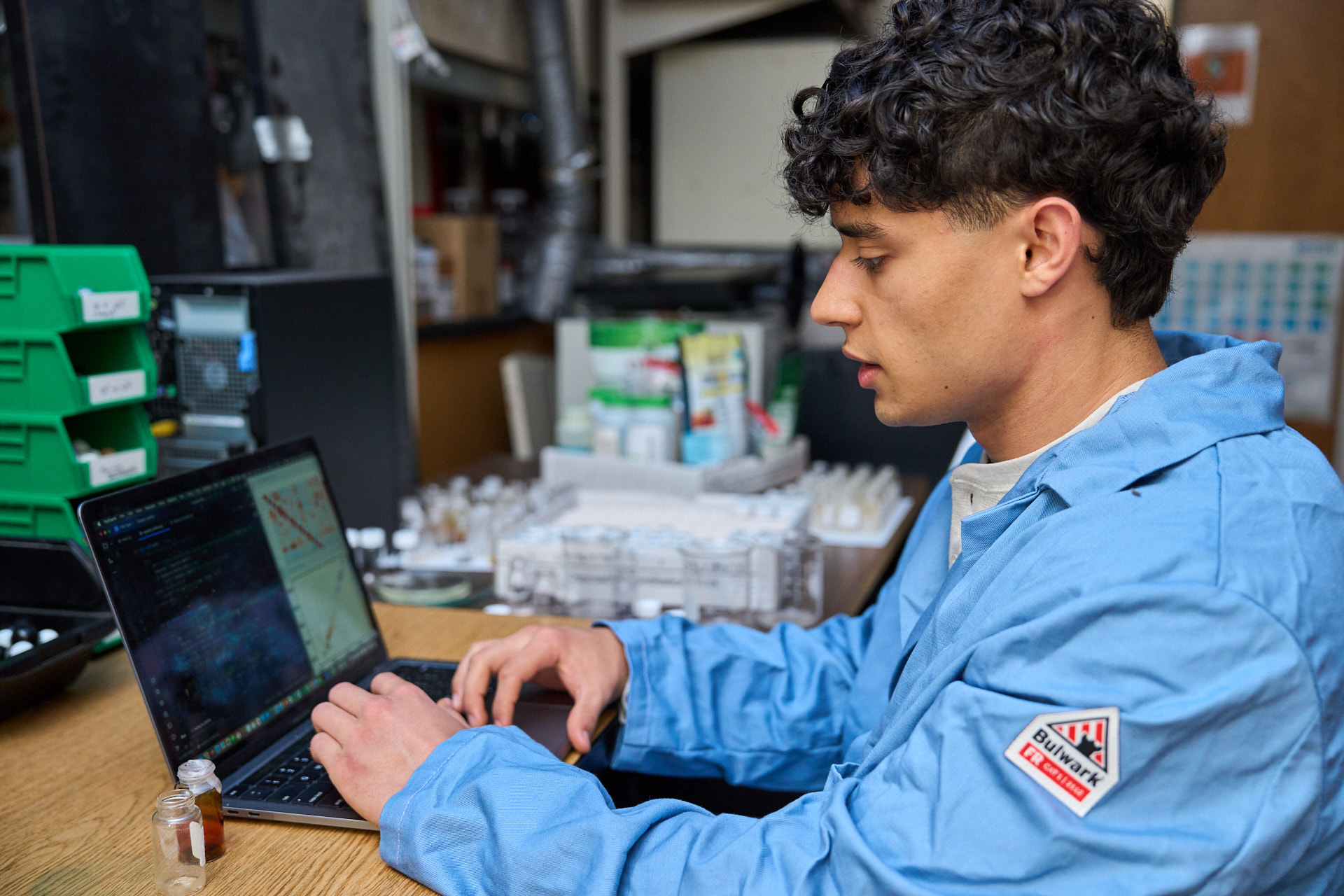 A young man wearing a blue lab coat types something into a laptop in a science lab.