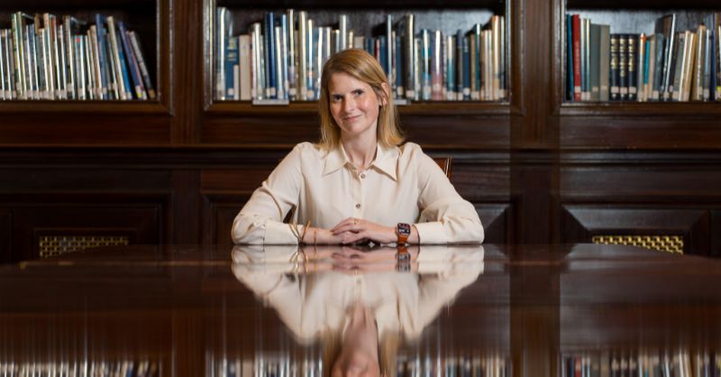 A woman sits at a table with books behind her