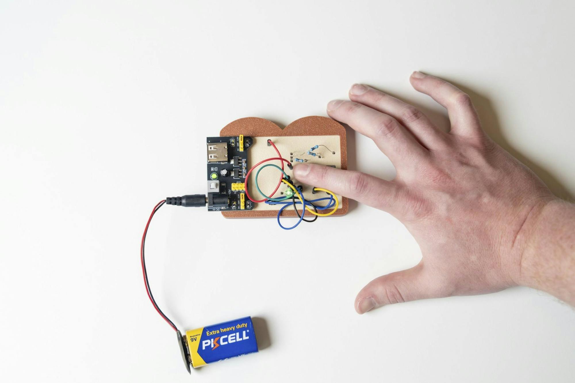 A hand rests on a circuit board with colorful wires, connected to a 9-volt battery lying nearby.
