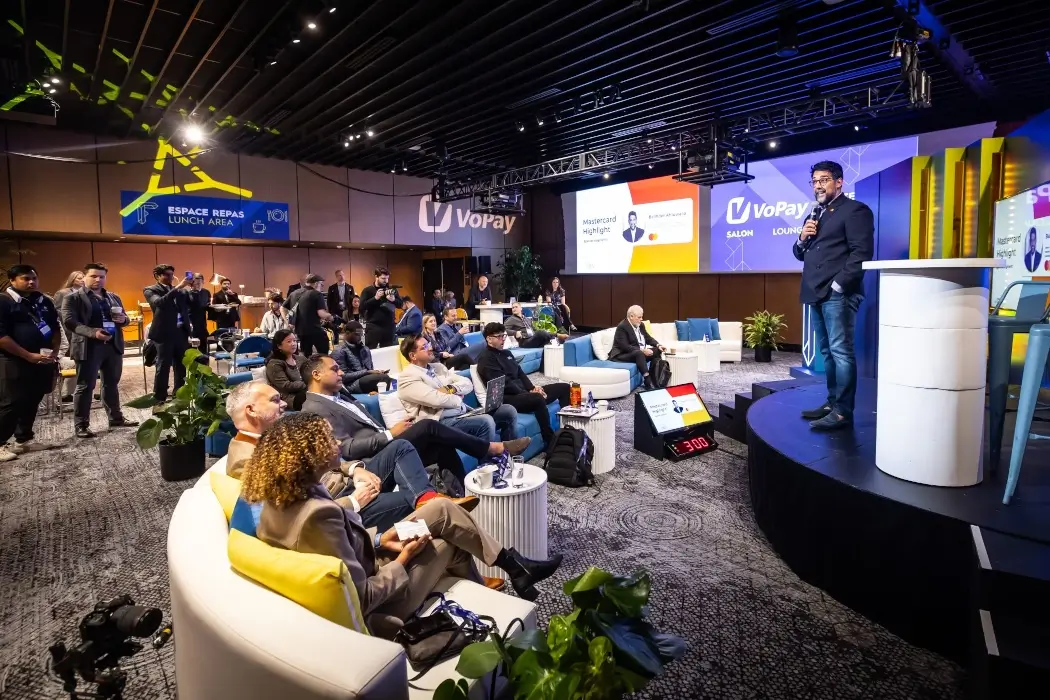 A large room with comfy-looking chairs and a stage, filled with people listening to a speaker.