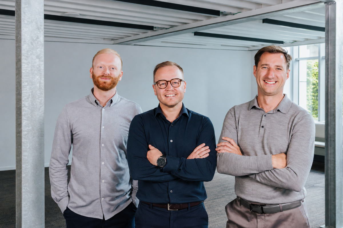 Three smiling male founding team members in business casual attire standing together indoors.